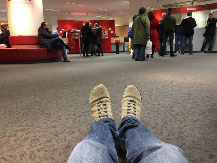 View from the floor of a bank branch over my own feet in light shoes and jeans, with customers queuing at the counter in the background