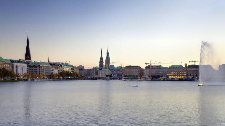 View across the Inner Alster Lake to Hamburg’s city centre with church spires and the Alster fountain at dusk.