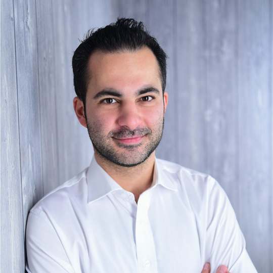 Hamed Farhadian im weißen Hemd steht mit verschränkten Armen vor einer grauen Wand und lächelt.