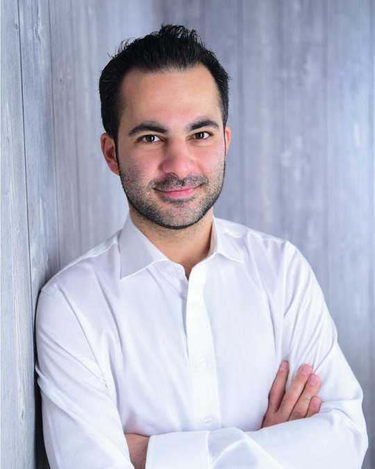 Hamed Farhadian im weißen Hemd steht mit verschränkten Armen vor einer grauen Wand und lächelt.