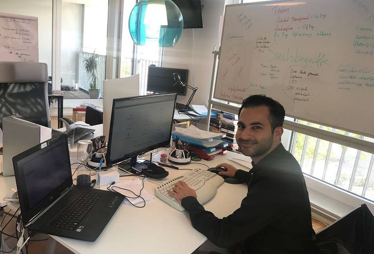 Hamed Farhadian am Schreibtisch im Büro, vor zwei Bildschirmen und einem vollgeschriebenen Whiteboard